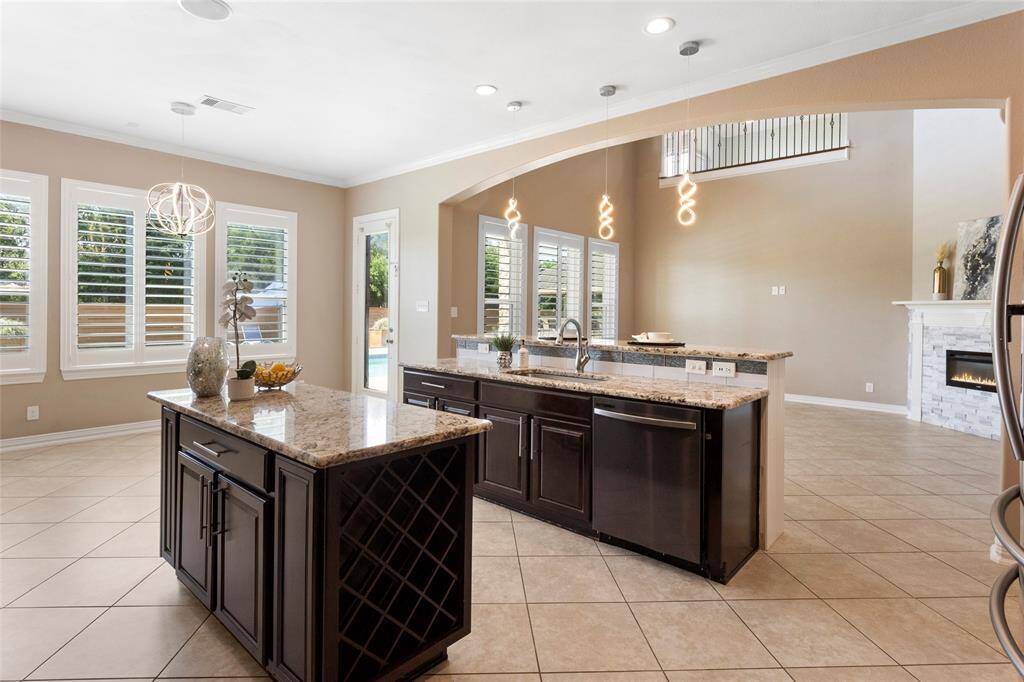 The kitchen features a large island with granite countertops, dark cabinetry, and modern pendant lighting. Multiple windows with plantation shutters allow for plenty of natural light, and a beautiful view of the backyard. The living area includes tiled flooring and a cozy fireplace, creating an inviting atmosphere.