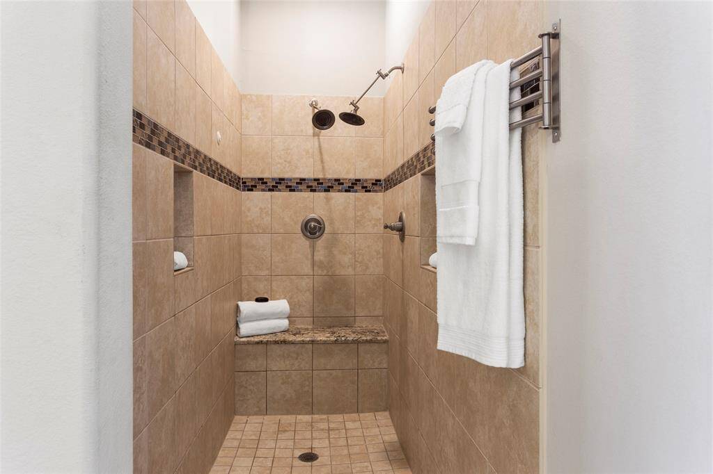 Check out this spacious, 7-foot walk-in shower with tile walls and flooring, accented by a decorative tile strip. It features dual shower heads, a built-in bench, and a towel rack, creating a functional and elegant bathroom space.