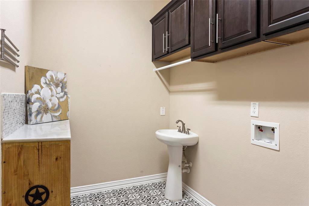 This laundry room features patterned tile flooring, a pedestal sink, dark wood cabinets for storage, and hookups for a washer and dryer.