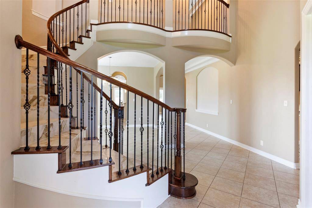 The elegant foyer leads upstairs with a sweeping staircase featuring wrought iron railings and marble steps. The open layout includes arched doorways and tile flooring, creating a welcoming and sophisticated entrance.