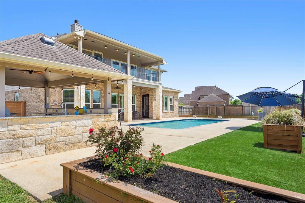 This photo showcases a spacious backyard with a swimming pool and a covered patio area, perfect for outdoor entertaining. The house features a two-story design with a balcony overlooking the pool. The yard is landscaped with planters and artificial turf.