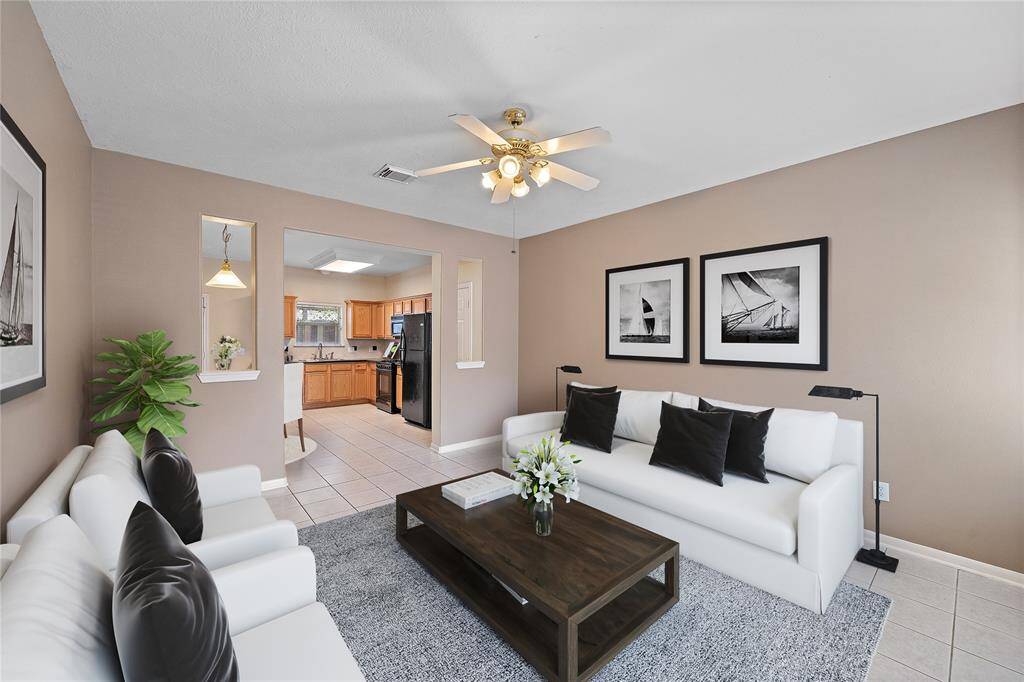 This virtually staged photo features a bright, open-concept living room with neutral walls and tile flooring. The space flows into a kitchen with wooden cabinets and modern appliances, creating a welcoming and functional living area.