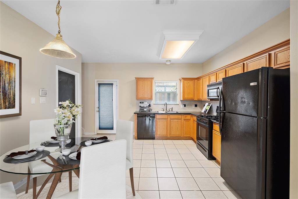 This virtually staged photo showcases a bright kitchen with wooden cabinets, black appliances, and a tiled floor. (NO CARPET THROUGHOUT HOME) The room has a modern, clean look with neutral walls and a decorative wall piece. The kitchen has easy access to the attached garage, as well as backyard patio.