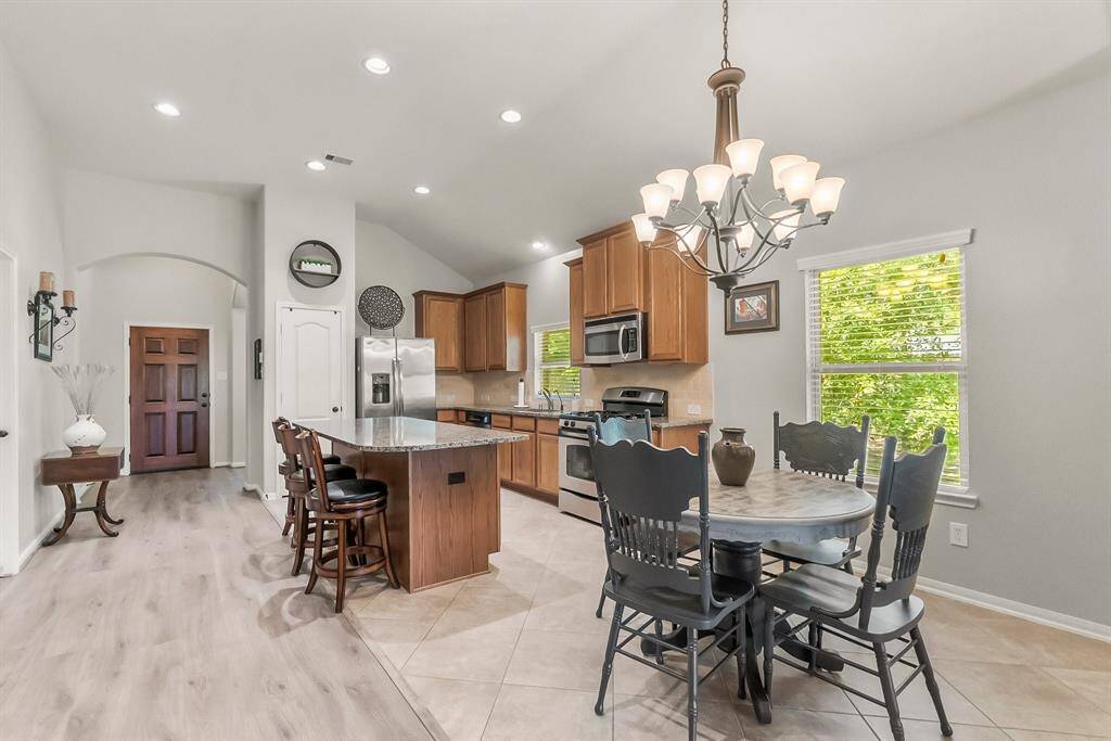Bright, open kitchen and dining area with modern appliances, wooden cabinets, and a center island with seating. The space features a stylish chandelier, large windows providing natural light, and light vinyl flooring that leads to the front entrance.