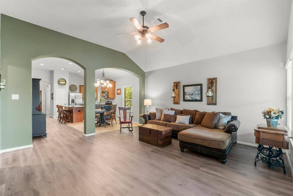 This inviting living space features an open floor plan with a high ceiling and warm wood flooring. The room is brightly lit, showcasing a cozy seating area and a dining space adjacent to the kitchen, enhanced by decorative arches and a ceiling fan.