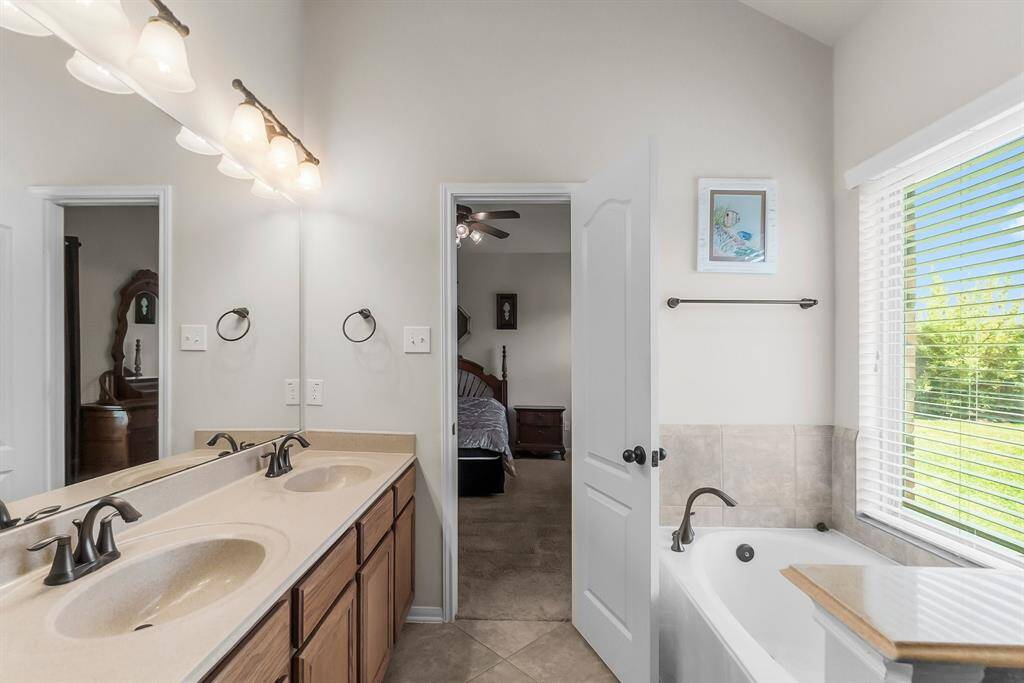 A bright bathroom featuring a double-sink vanity with ample lighting, a soaking tub with a view of the outdoors, and an open doorway leading to the primary bedroom.
