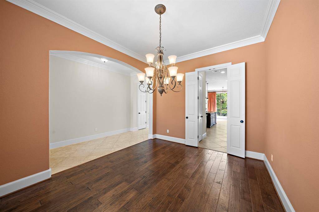 This photo showcases a bright dining room with warm orange walls, elegant crown molding, and rich hardwood floors. A stylish chandelier centers the space, and double doors open to a view of the kitchen and living area, enhancing openness and flow.