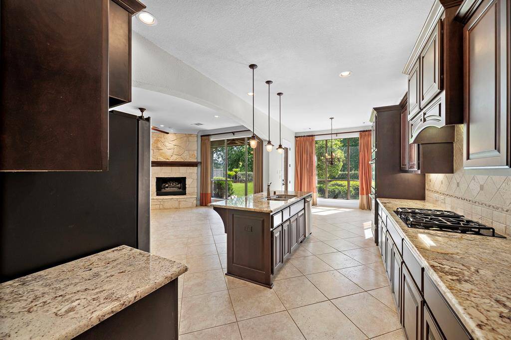 This spacious kitchen features granite countertops, dark wood cabinets, and a central island with pendant lighting. It opens to a cozy living area with a stone fireplace and large windows that offer views of the lush backyard.