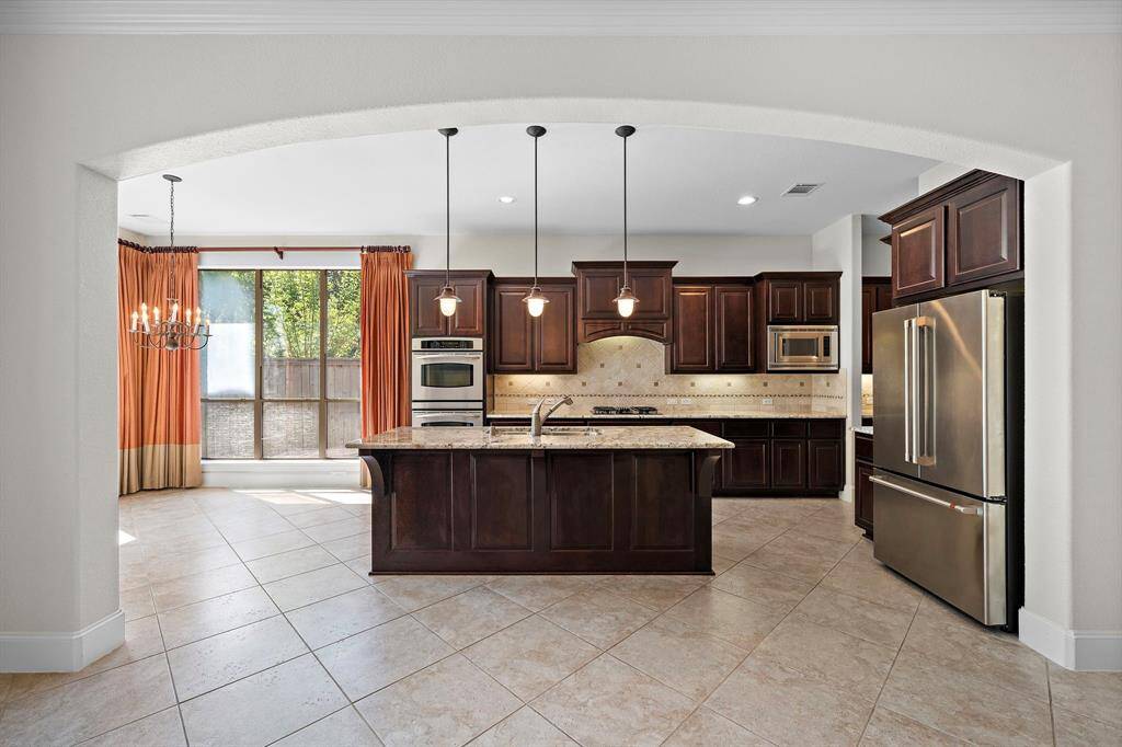 Elegant kitchen with dark wood cabinetry, granite countertops, and stainless steel appliances. Features a central island, pendant lighting, and a large window with orange drapes, providing plenty of natural light.