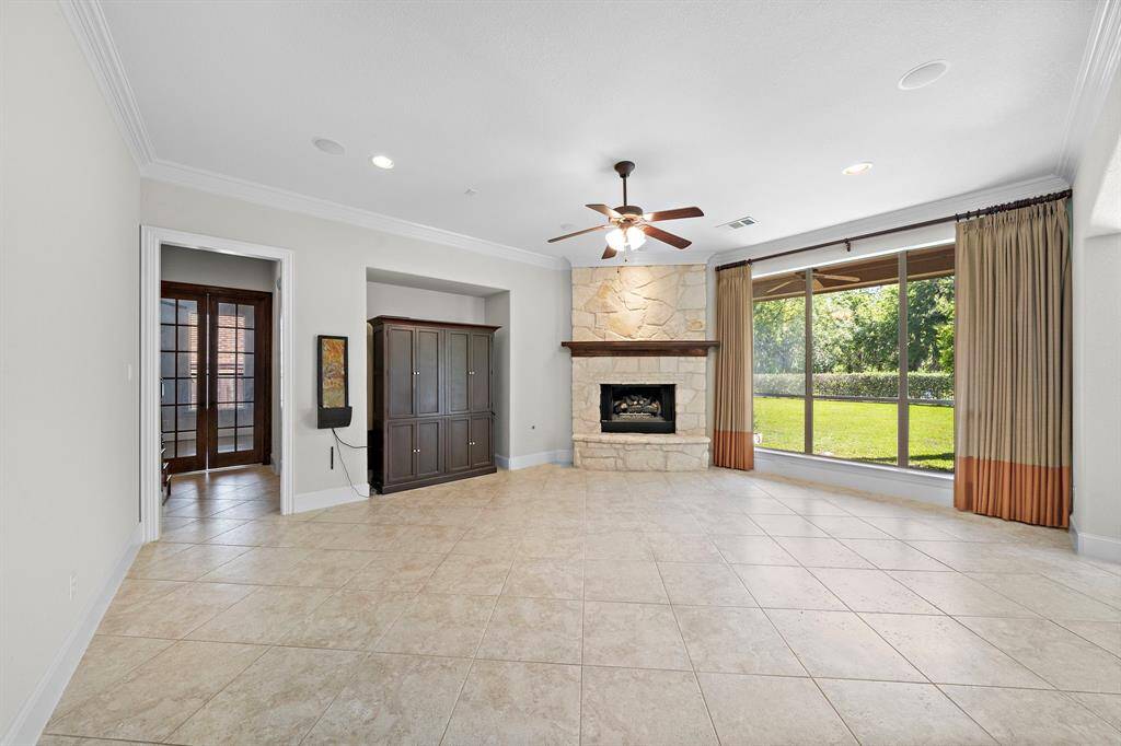 This spacious living room features large tiled flooring, a stone fireplace, and a ceiling fan. It has ample natural light from expansive windows overlooking a lush backyard. There's a built-in entertainment unit, and French doors lead to an adjacent room.