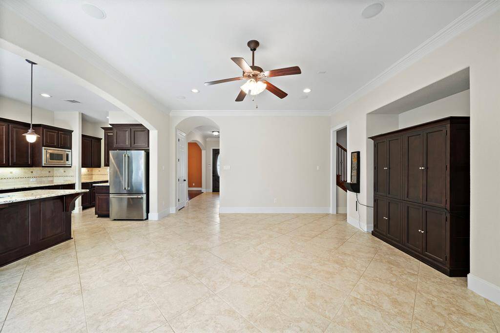 This photo features a spacious, open-concept living area with tiled floors, a ceiling fan, and neutral walls. It connects to a modern kitchen with dark wood cabinetry, stainless steel appliances, and a breakfast bar. The room is bright with ample natural light.