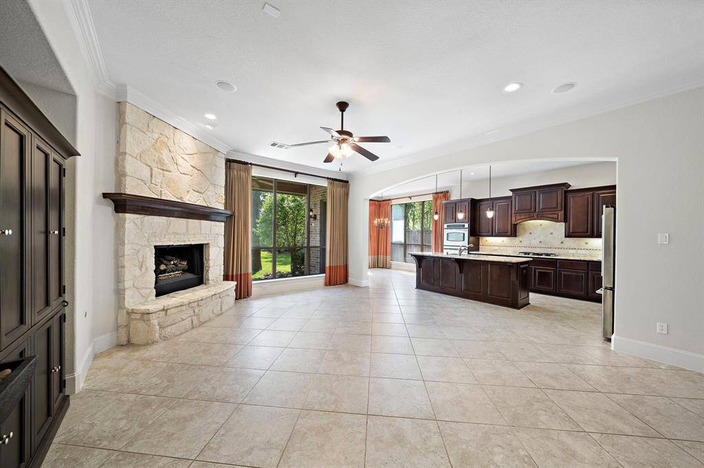 This spacious living area features a stone fireplace, large windows with garden views, and an open layout leading to a modern kitchen with dark wood cabinetry and stainless steel appliances. The room is bright and airy, with tile flooring and a ceiling fan.