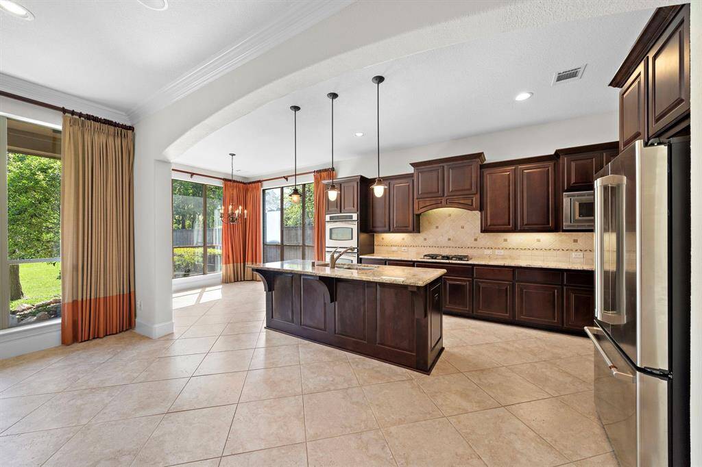 This spacious kitchen features elegant dark wood cabinets, granite countertops, and stainless steel appliances. An island with pendant lighting offers extra prep space. Large windows and an adjacent dining area with a chandelier provide abundant natural light and a view of the greenery outside.