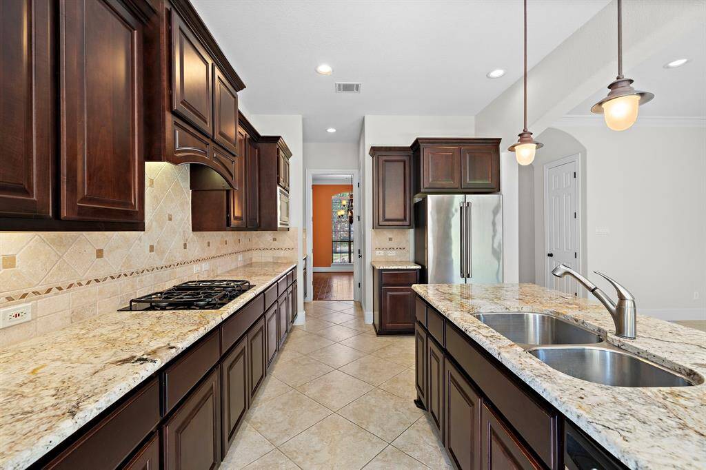 This kitchen features elegant dark wood cabinetry, granite countertops, and a stainless steel refrigerator. It includes a gas cooktop, double sink, and pendant lighting, providing a spacious and modern cooking area with a tiled backsplash and floor.