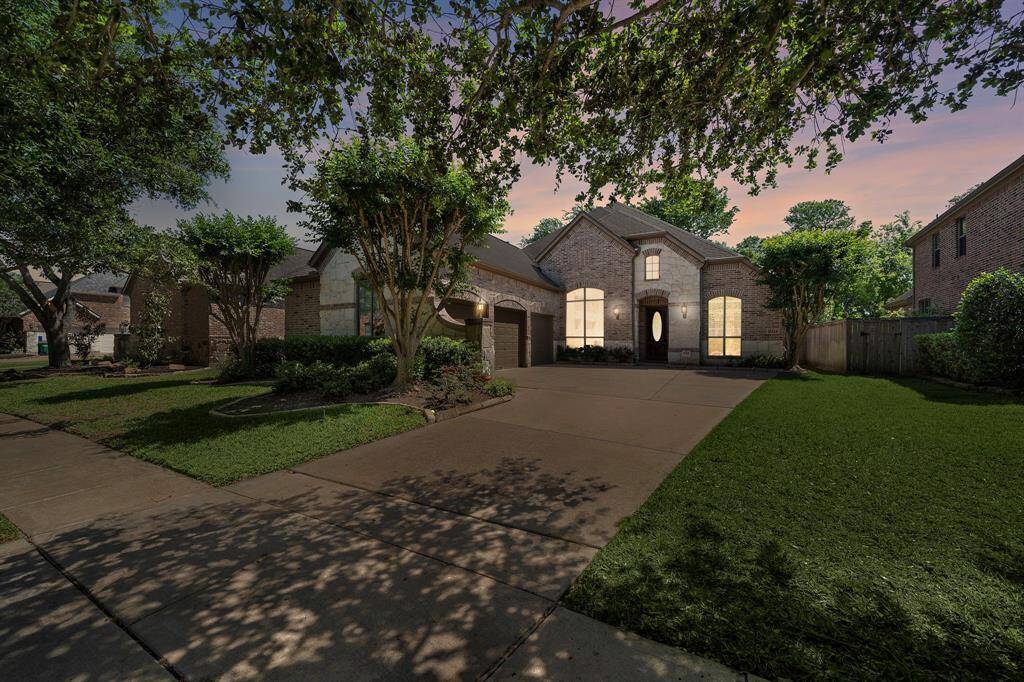 This photo showcases a charming two-story brick home with a well-maintained front yard and mature trees providing shade. The driveway leads to a two-car garage, and the house features large, warmly lit windows, creating an inviting appearance at dusk.