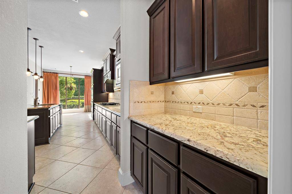This photo showcases a modern kitchen with sleek dark cabinetry, light granite countertops, and elegant tile backsplash. The space is brightened by natural light from large windows, and features pendant lighting and a spacious layout with tile flooring.