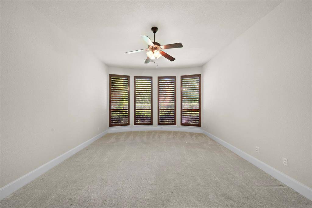 This room features neutral walls and carpet, a ceiling fan with lights, and large windows with wooden shutters, providing ample natural light.