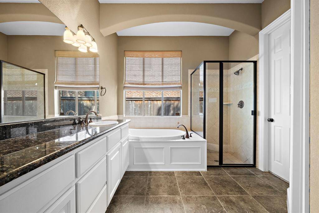 This bathroom features a spacious layout with a large granite countertop, dual sinks, a soaking tub, and a glass-enclosed shower. The neutral-toned tiles and natural light create a warm and inviting atmosphere.
