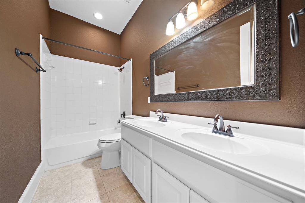 This bathroom features a double sink vanity with a large, decorative mirror and modern lighting. It has a bathtub-shower combo with white tile surround, complemented by warm brown walls and beige flooring.