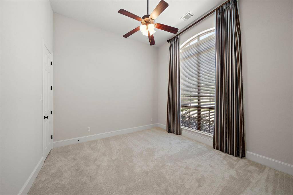 This room features a neutral color palette with carpeted flooring, a ceiling fan, and a large window with elegant drapes, allowing plenty of natural light. Ideal for a cozy bedroom or home office.