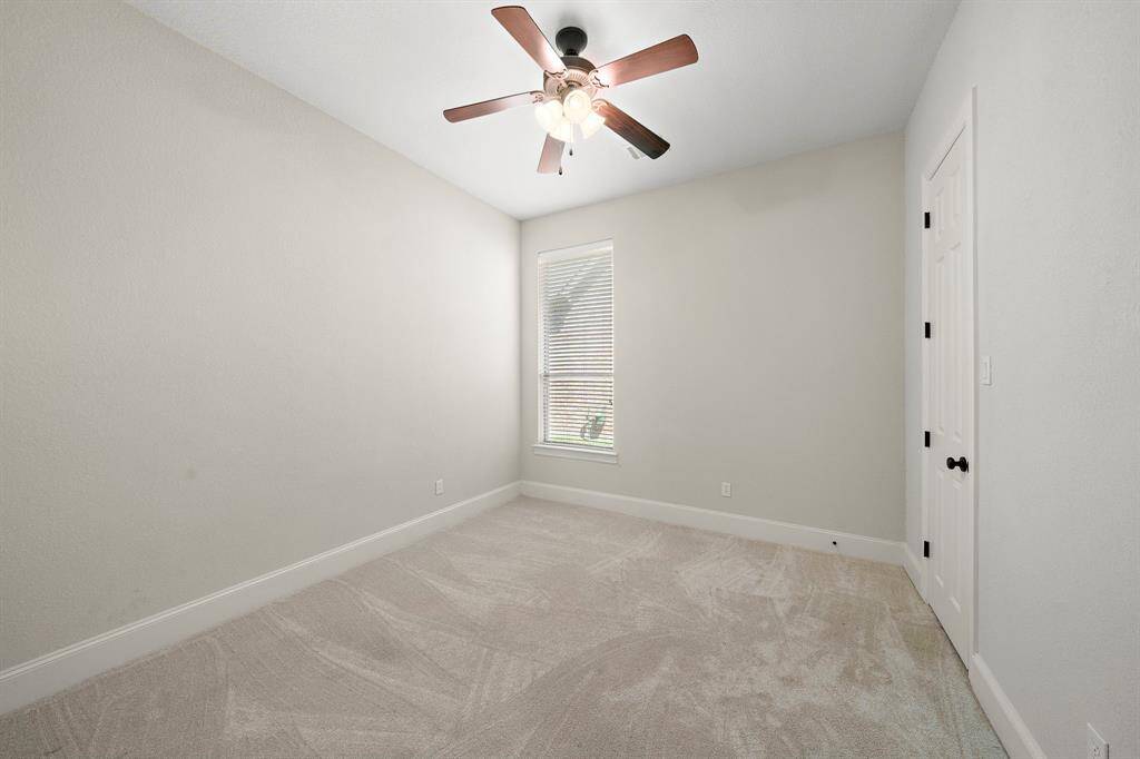 This is a bright, small bedroom with neutral walls and carpet. It features a ceiling fan and a single window that lets in natural light, creating a cozy atmosphere.