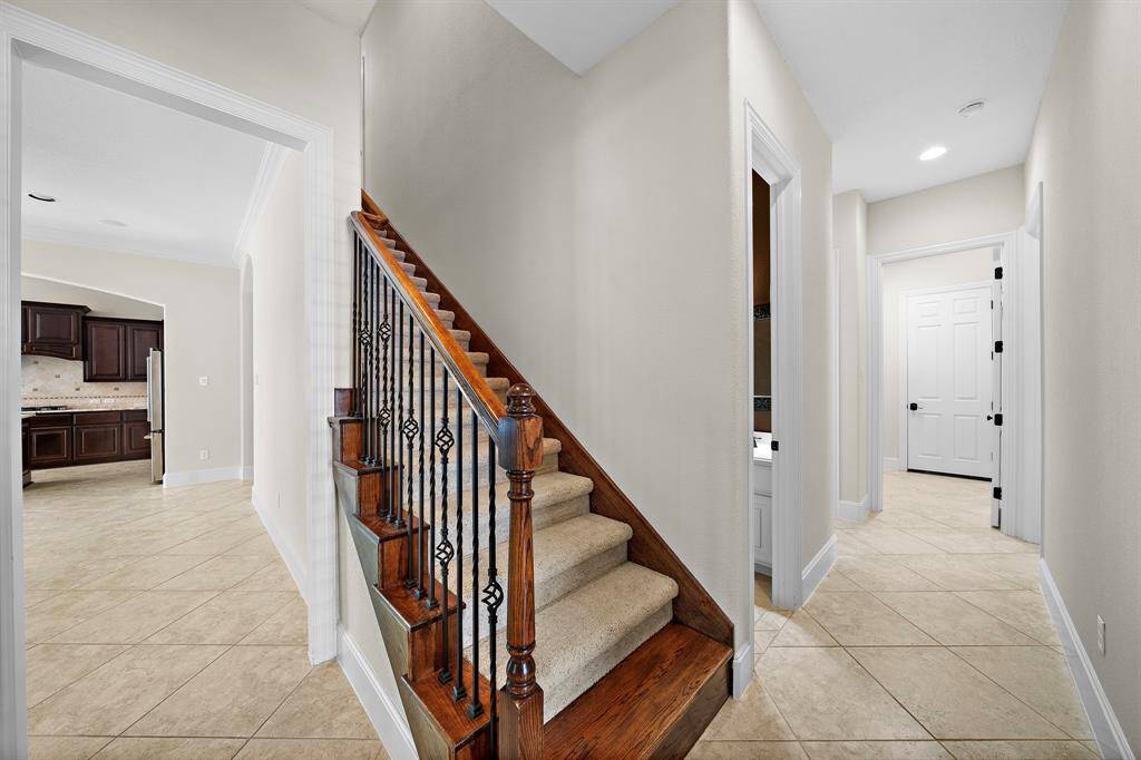 This photo showcases a bright and spacious hallway with tiled floors and a carpeted staircase featuring elegant wooden railings. The hallway leads to a kitchen with dark cabinetry and modern finishes, offering a warm and inviting atmosphere for a potential home buyer.