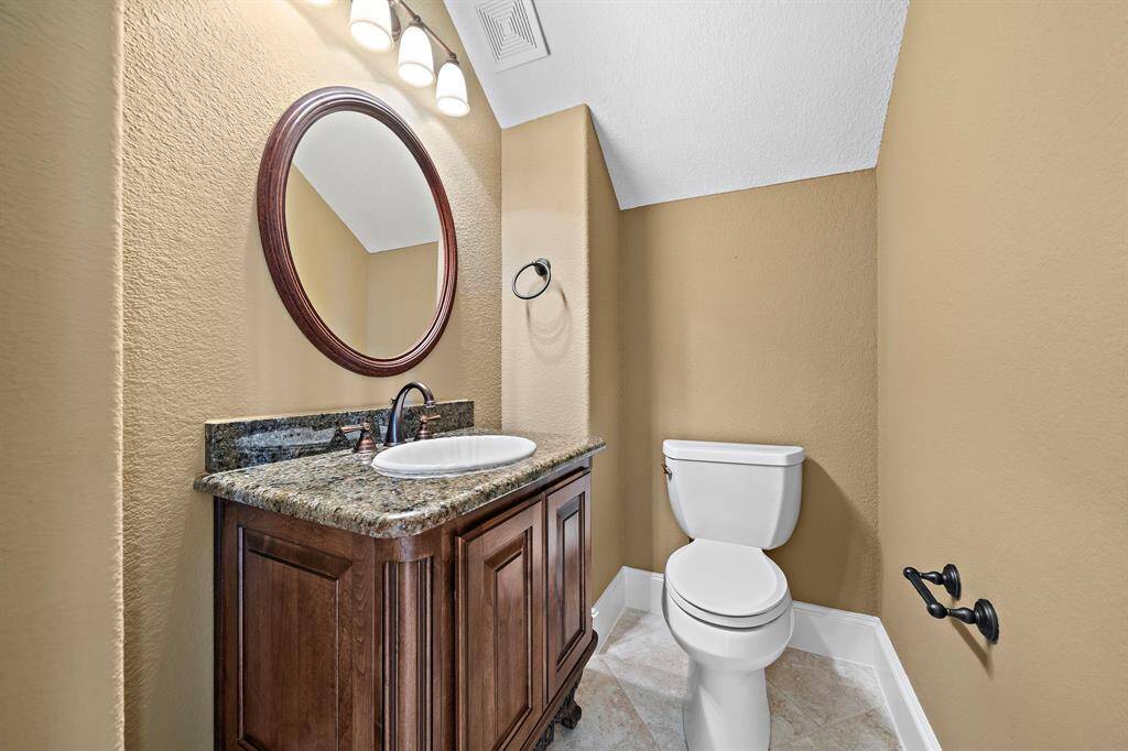 This is a cozy half-bathroom with beige walls featuring a classic wooden vanity with a granite countertop and undermount sink. It includes a round mirror and a standard toilet, with elegant lighting above.