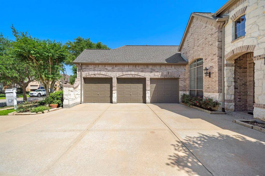 This photo showcases a spacious driveway leading to a three-car garage attached to a brick and stone home. The exterior features large windows and well-maintained landscaping, creating an inviting entrance.