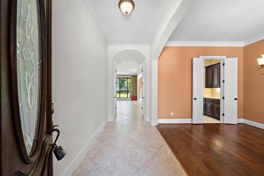 This photo showcases a welcoming entryway with a mix of tile and wood flooring. It features an arched doorway leading to a bright room with large windows overlooking greenery. To the right is a room with double doors and a view into a modern kitchen area.