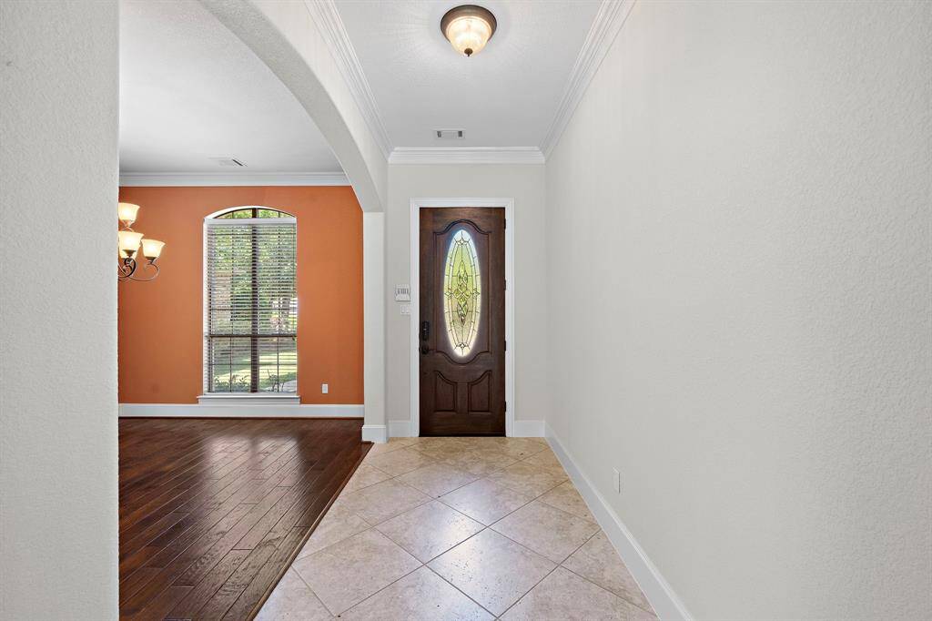 This photo showcases a welcoming entryway featuring a stylish wooden front door with a decorative glass panel. The space has a neutral tile floor leading into a room with warm, orange accent walls and rich hardwood flooring. Natural light streams through a large window, highlighting the elegant archway and crown molding.
