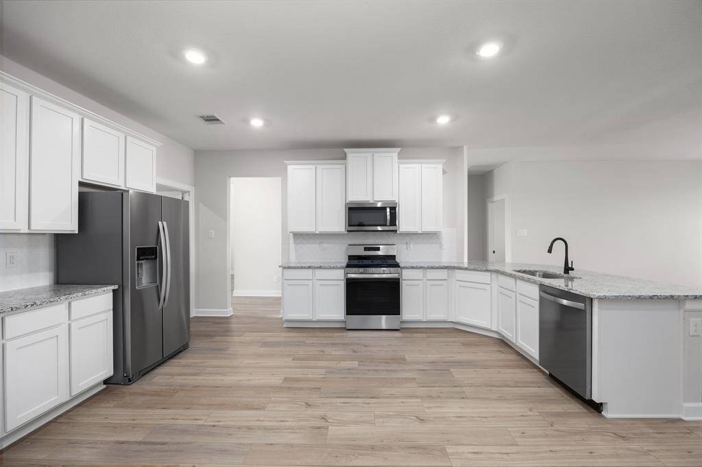 Spacious and modern kitchen featuring elegant white cabinetry, sleek granite countertops, and stainless steel appliances. The open layout includes a large breakfast bar with a built-in sink and stylish black faucet, perfect for entertaining. Beautiful wood-look flooring and a crisp white subway tile backsplash complete the clean, contemporary design.