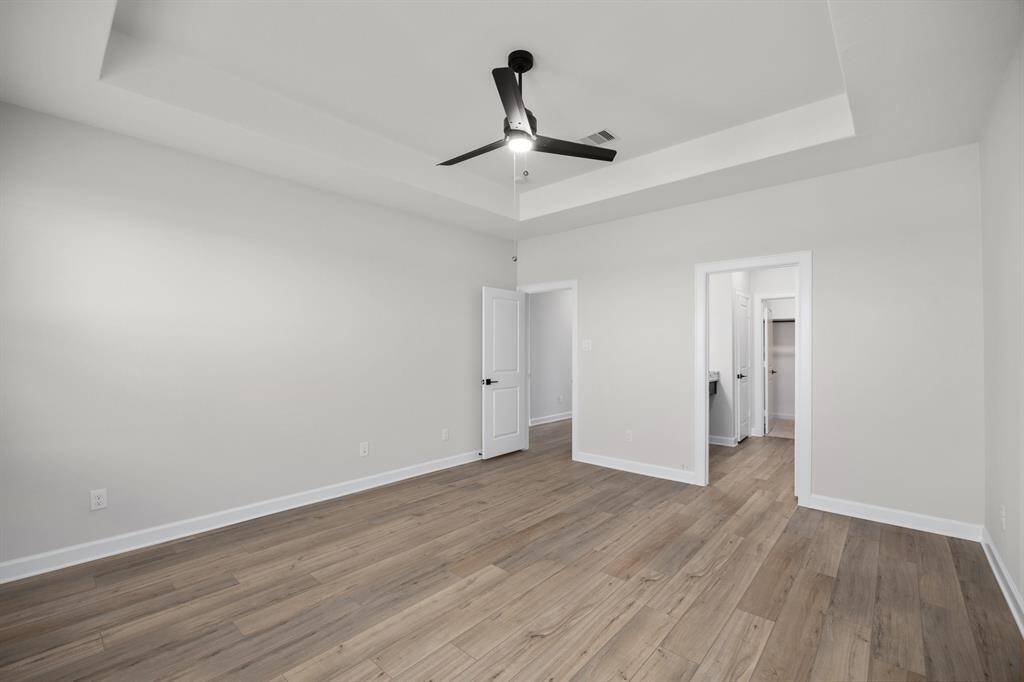 Bright and airy primary bedroom featuring a tray ceiling with a modern ceiling fan, large windows for ample natural light, and stylish wood-look flooring. The neutral color palette provides a serene retreat, ready for your personal touch.