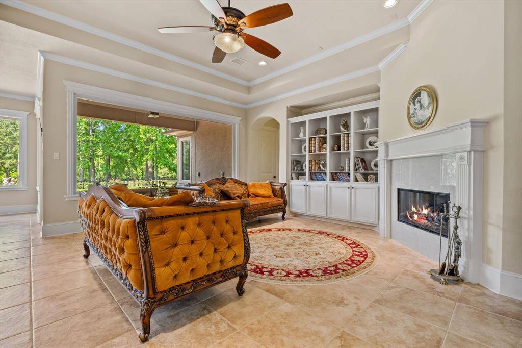 This elegant living room features a cozy fireplace, built-in shelves, and classic furniture with ornate details. The space is well-lit with recessed lighting and a ceiling fan, and it opens to a hallway with arched doorways, creating a warm and welcoming atmosphere.