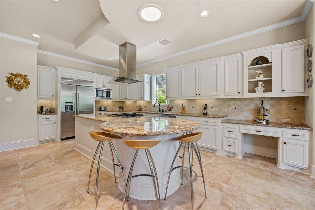 This spacious kitchen features elegant white cabinetry, a large center island with a granite countertop, and modern stainless steel appliances. The open layout flows into a bright dining area with a large window overlooking a lush backyard, perfect for entertaining and family gatherings.