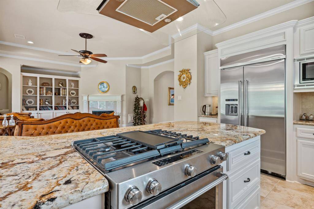 This spacious kitchen features elegant granite countertops, a stainless steel gas stove, and a double-door refrigerator. It opens into a cozy living area with built-in shelves and a stylish ceiling fan, offering a great space for entertaining.