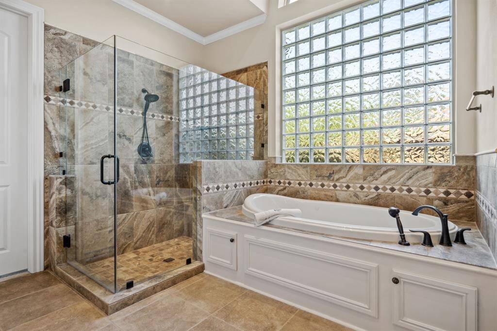 Indulge in spa-like relaxation in this luxurious primary bath, featuring a walk-in glass shower, deep soaking tub, and elegant tile finishes throughout. Natural light filters in through the glass block window, creating a serene and private retreat.