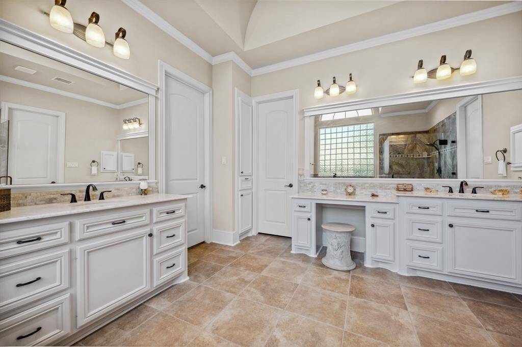 This spacious bathroom features dual sinks with elegant fixtures, ample cabinetry, a makeup vanity, and large mirrors. It has a warm, neutral color palette and is well-lit, creating a luxurious and inviting atmosphere.