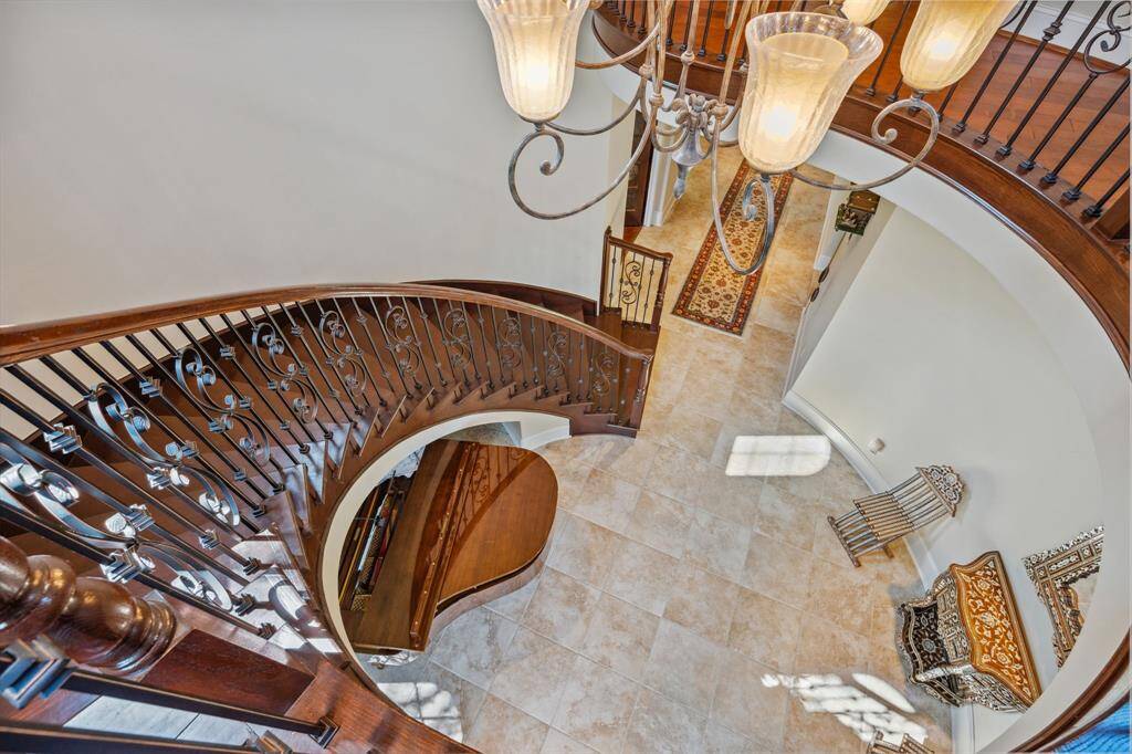This breathtaking view from the second floor showcases the graceful curve of the staircase, intricate wrought iron railing, and elegant chandelier lighting the grand foyer below. A statement of timeless architecture and refined design, this entryway leaves a lasting impression.