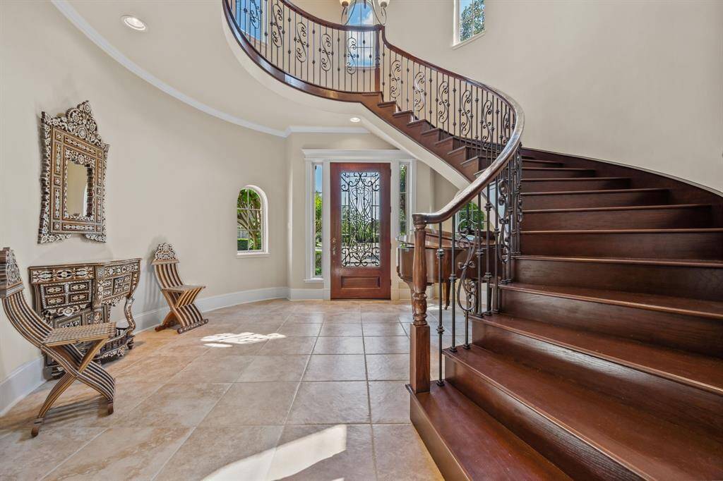 Step into elegance with this grand foyer featuring a dramatic sweeping staircase, rich wood accents, and wrought iron detailing. Natural light pours through the custom front door and surrounding windows, creating a warm and welcoming first impression.