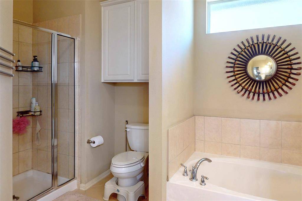 Another view of the primary bathroom features a spacious walk-in shower with glass doors, a bathtub with modern fixtures, a toilet, and a decorative circular mirror. It has a clean, neutral color palette with ample natural light.