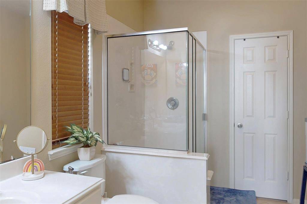 This first floor ensuite bathroom of the first floor bedroom features a modern glass-enclosed shower, a vanity with a round mirror, and a window with wooden blinds. The space is bright and clean, and complemented by neutral tones