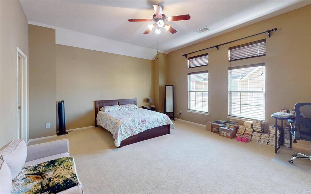 This spacious primary bedroom features neutral walls, large windows with ample natural light, a ceiling fan, and a cozy carpeted floor. There's room for a bed, seating area, and workspace, creating a comfortable and versatile living space.