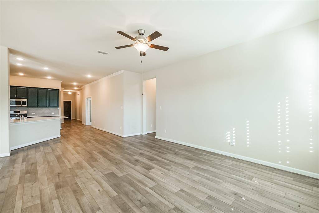 This spacious living area features wood flooring, neutral walls, and a ceiling fan. The open layout connects seamlessly to a modern kitchen with dark cabinetry and stainless steel appliances, ideal for entertaining and family gatherings.