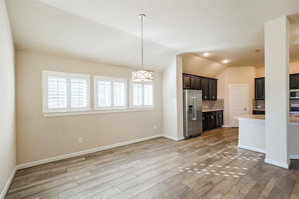Bright, open-concept space with light wood flooring and neutral walls. Features a modern kitchen with dark cabinets, stainless steel appliances, and a breakfast bar. Natural light streams through windows with plantation shutters, enhancing the airy feel.