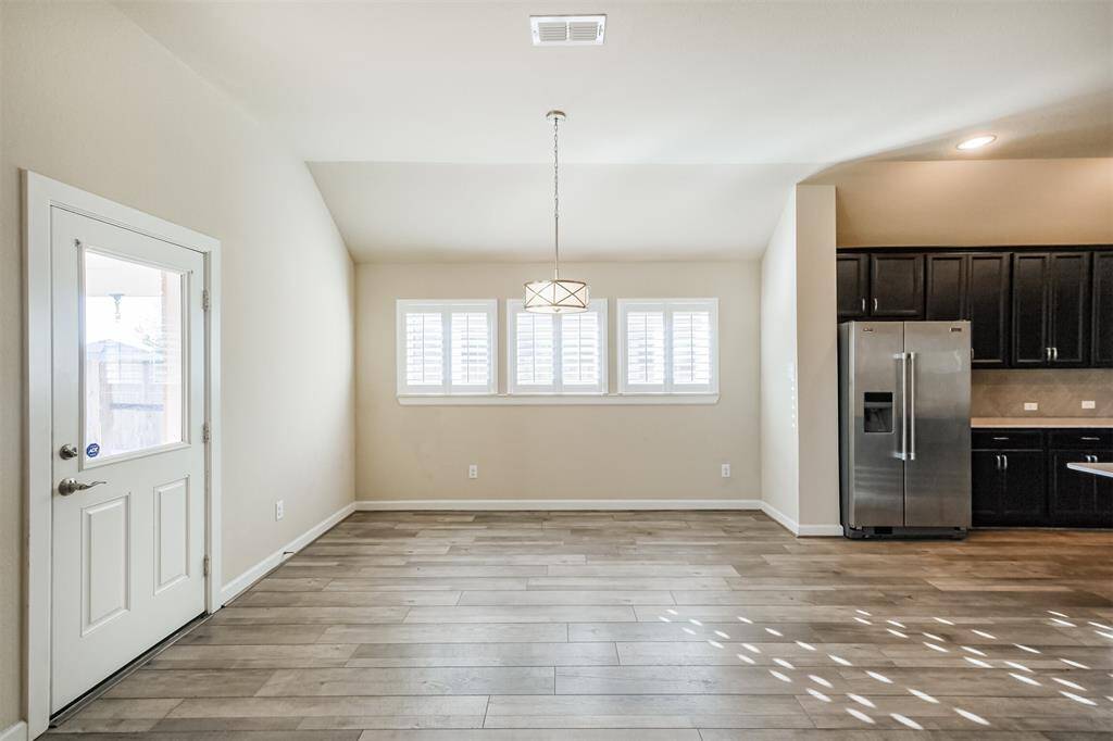 This bright dining area features large windows with wooden shutters, a modern light fixture, and wood-look flooring. It opens to a kitchen with dark cabinets and stainless-steel appliances, creating a sleek and inviting space.