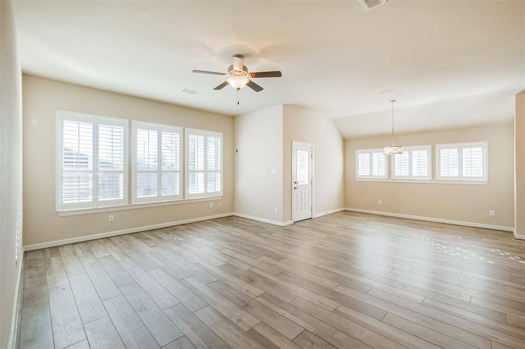 This spacious, bright living room features large windows with wooden shutters, light wood flooring, and a modern ceiling fan. The open layout includes a dining area with a stylish light fixture, creating a welcoming and airy atmosphere.