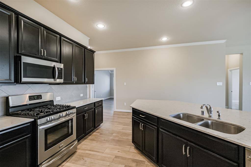 This kitchen features modern stainless steel appliances, dark cabinetry, and a spacious island with a double sink, all set against light-colored walls and wood flooring.