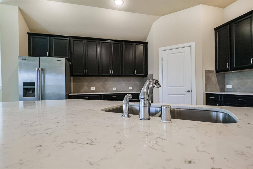 This kitchen features elegant dark cabinetry, a sleek stainless steel refrigerator, and a modern marble countertop with a built-in sink. The space is well-lit, offering a clean and contemporary feel.