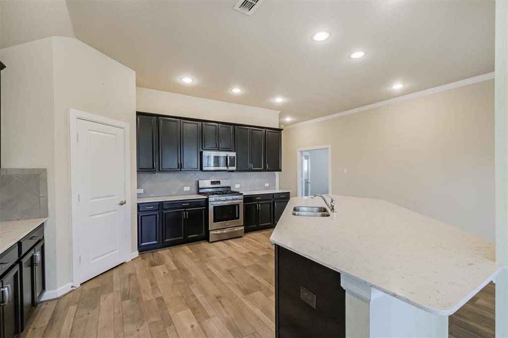 This modern kitchen features dark cabinetry, stainless steel appliances, and light countertops. It has recessed lighting, wood-style flooring, and a spacious layout with a central island sink.