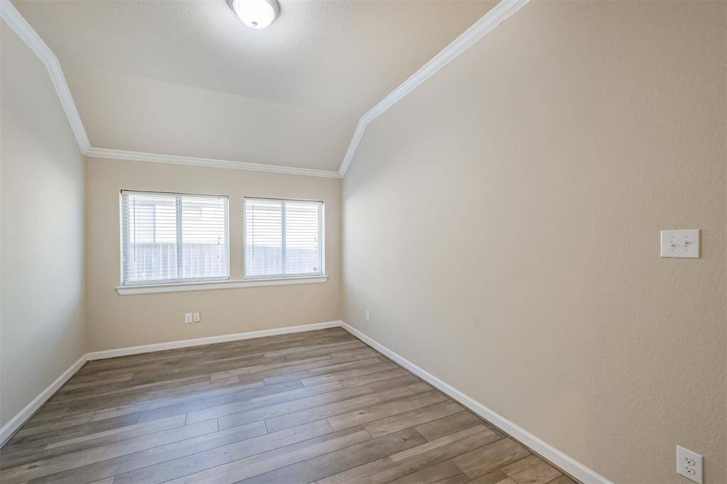 This primary bedroom features soft beige walls, wide plank wood flooring, and a vaulted ceiling with crown molding. Two large windows provide natural light, creating a bright and inviting space.